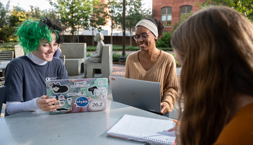 Three undergraduate students sit at a table on Georgia Tech’s campus, talking while studying around a notebook and two laptops.