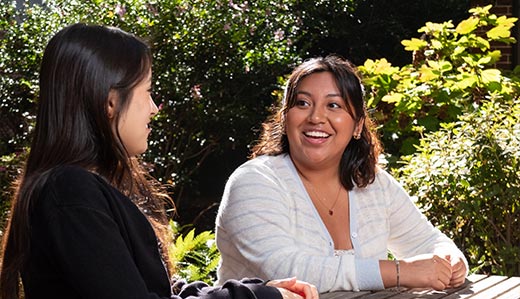 Two Tech Promise students sit at a wooden picnic table outside on Georgia Tech’s campus. The two students are having a conversation and smiling.